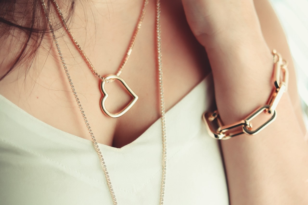 A close-up of a woman wearing layered yellow gold necklaces, one featuring an open heart pendant, paired with a chunky yellow gold chain bracelet.