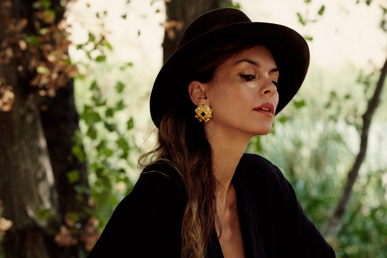 A woman in a dark outfit and wide-brimmed hat poses outdoors, wearing massive yellow gold floral statement earrings.