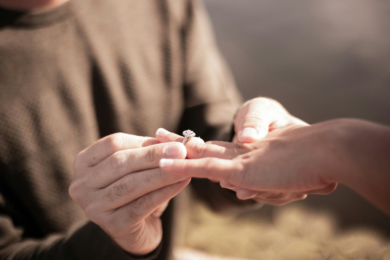 A man down on one knee putting a halo engagement ring on a woman’s finger