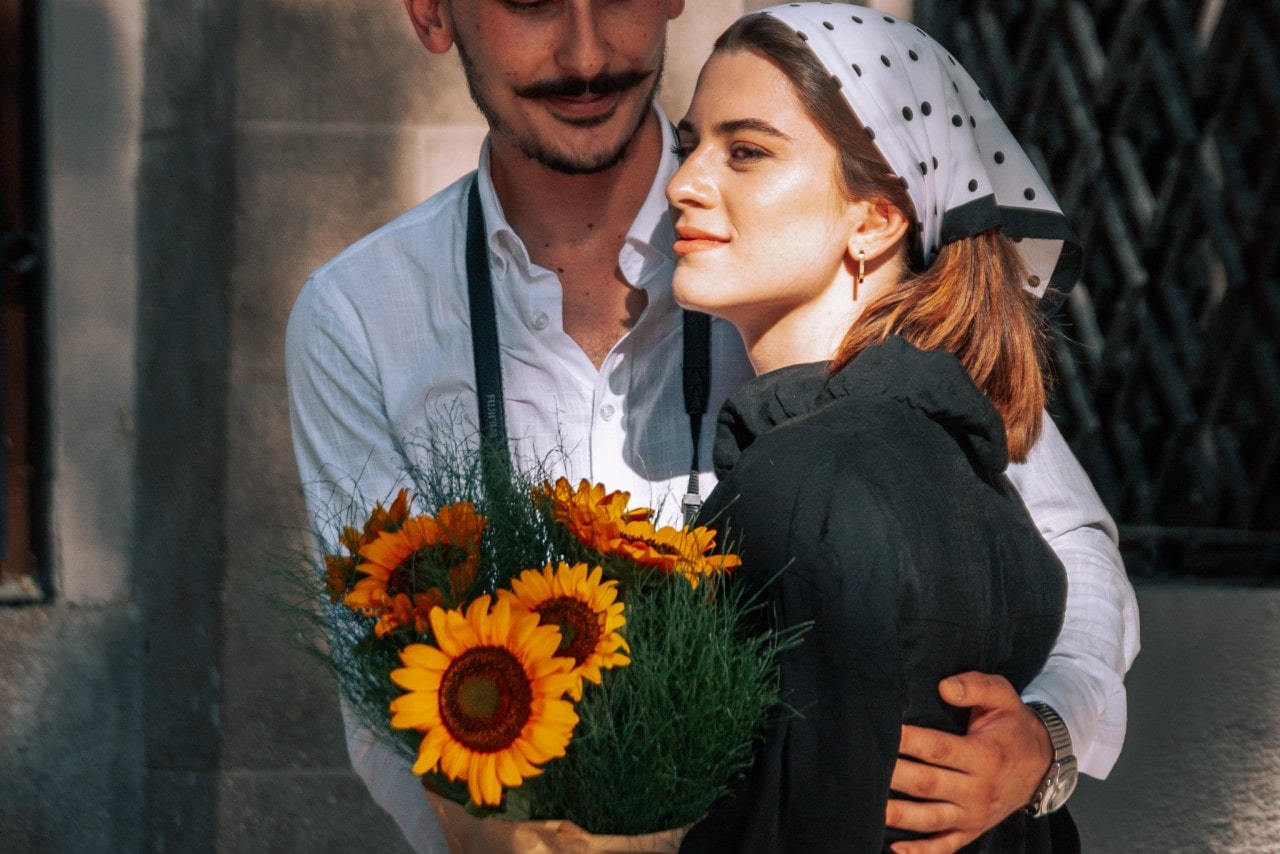 a couple embracing, the woman holding a bouquet of sunflowers and the man wearing a silver watch