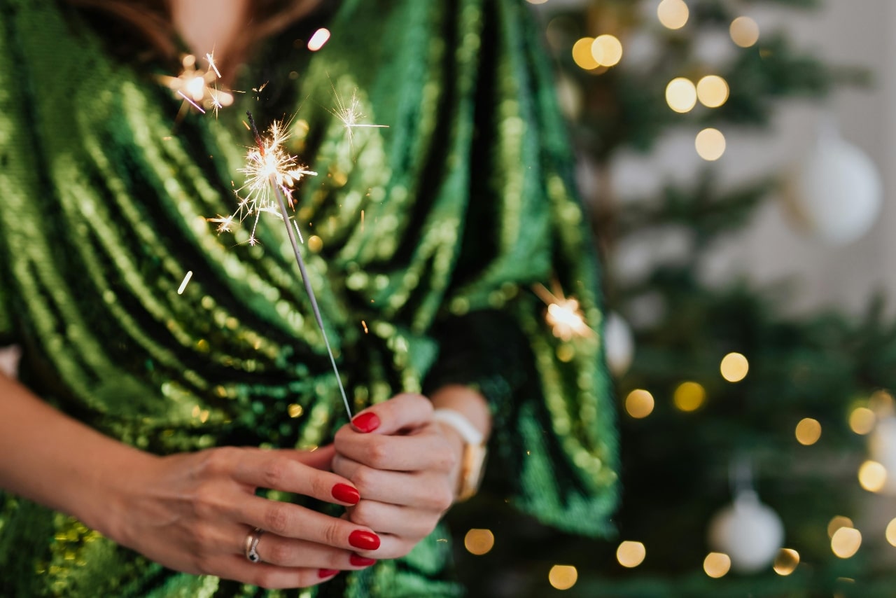 Woman in a shimmering green dress holding a lit sparkler in front of a blurred Christmas tree.