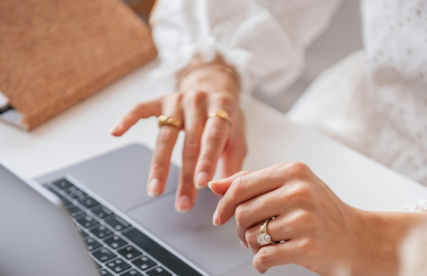 A close-up of a woman's hands adorned with gold and diamond rings as she types on a laptop, with a brown notebook in the background.
