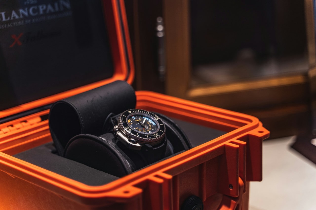A luxury watch in a vibrant orange protective case, displayed on black padding.