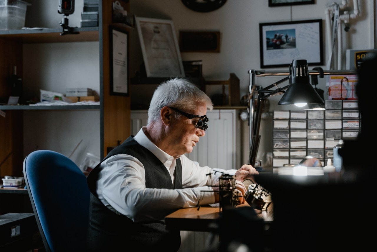 A man with gray hair wearing magnifying glasses works intently at a cluttered desk in a dimly lit workshop
