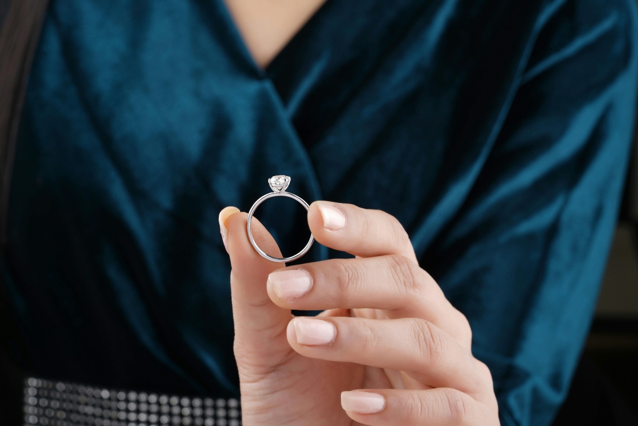 A close-up of a woman in a deep blue outfit holding a solitaire diamond engagement ring between fingers.