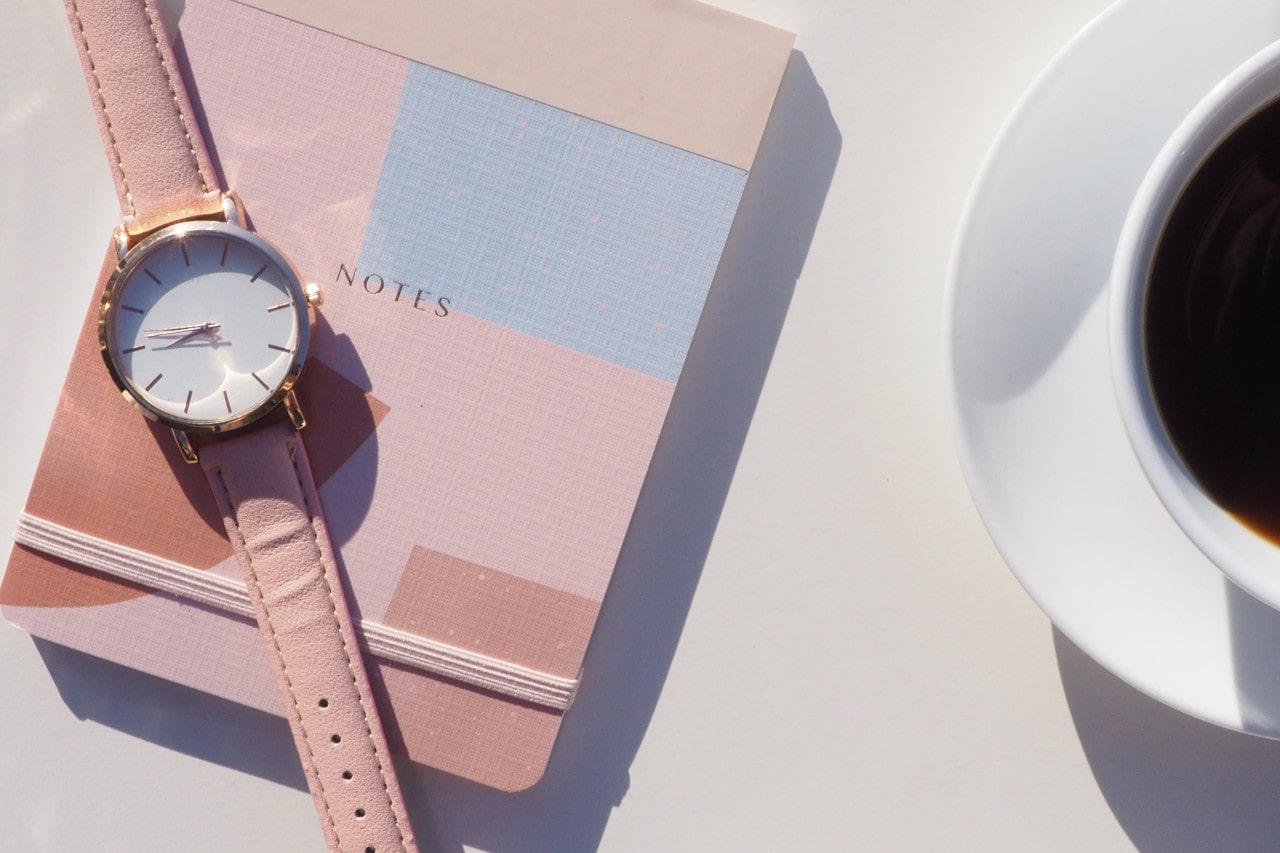 A pastel pink wristwatch rests on a pink notebook, accompanied by a cup of black coffee in bright natural light