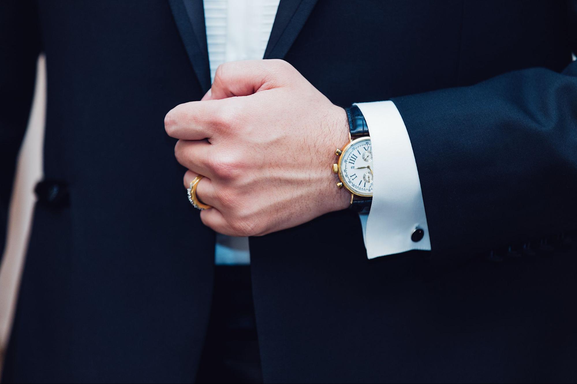 A man in a black suit wearing a gold wristwatch with a white face and blue pointers and a gold ring on his finger