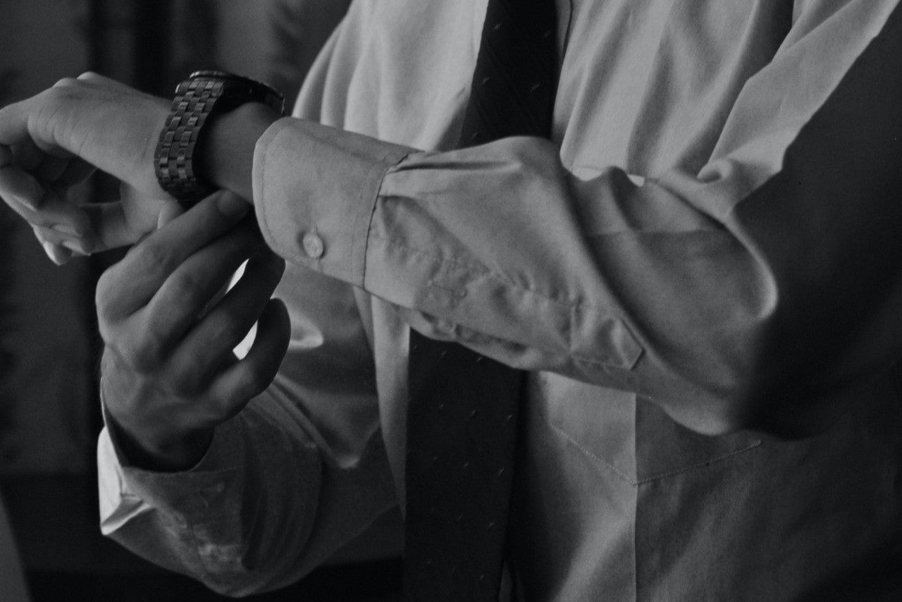 A black-and-white close-up of a man adjusting his metal wristwatch while wearing a dress shirt and tie