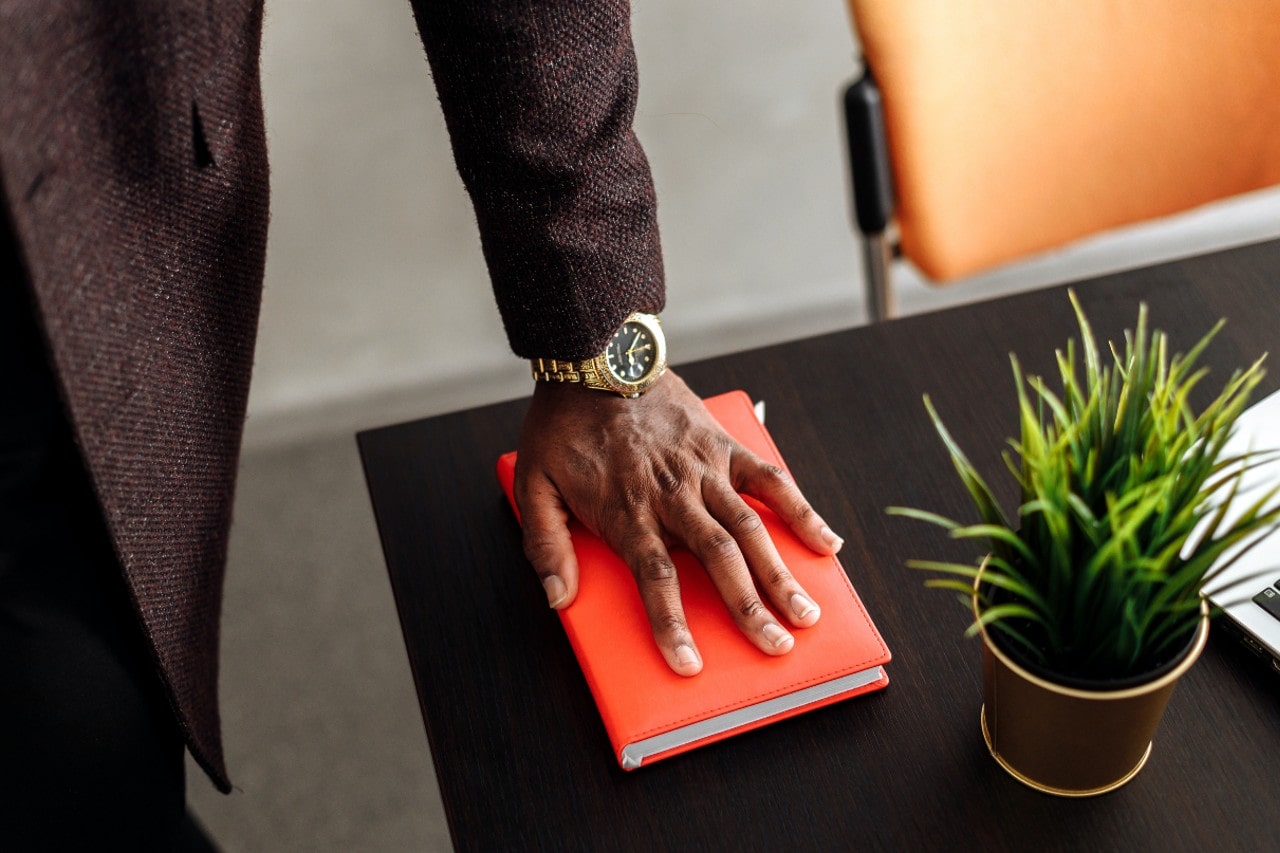A man in a dark blazer wears a gold wristwatch while placing his hand on an orange notebook on a sleek wooden desk with a potted plant