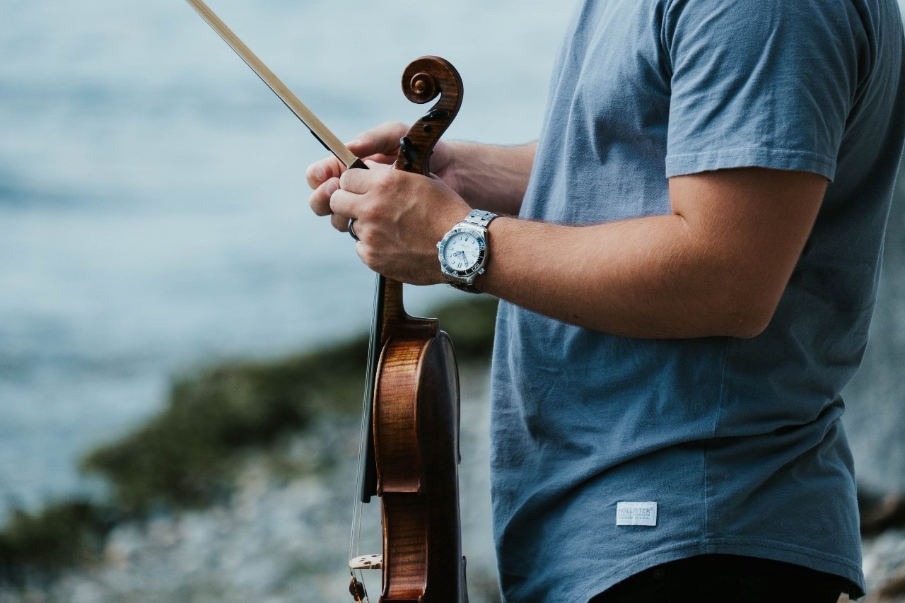 a man standing by the water, holding a violin and wearing a silver diver watch