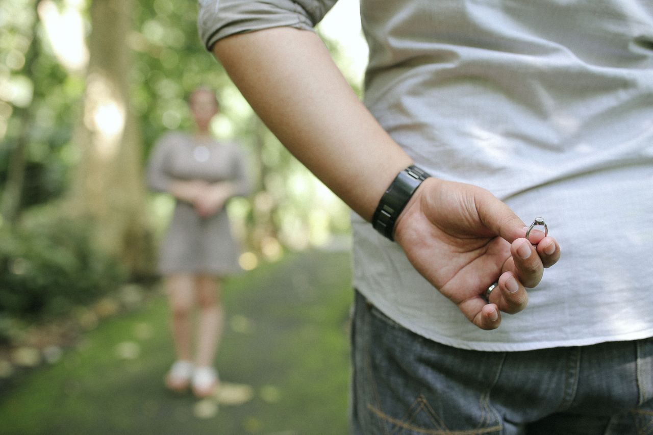 A man in casual attire stands on a lush green path, secretly holding an engagement ring behind his back while his partner waits in the distance.
