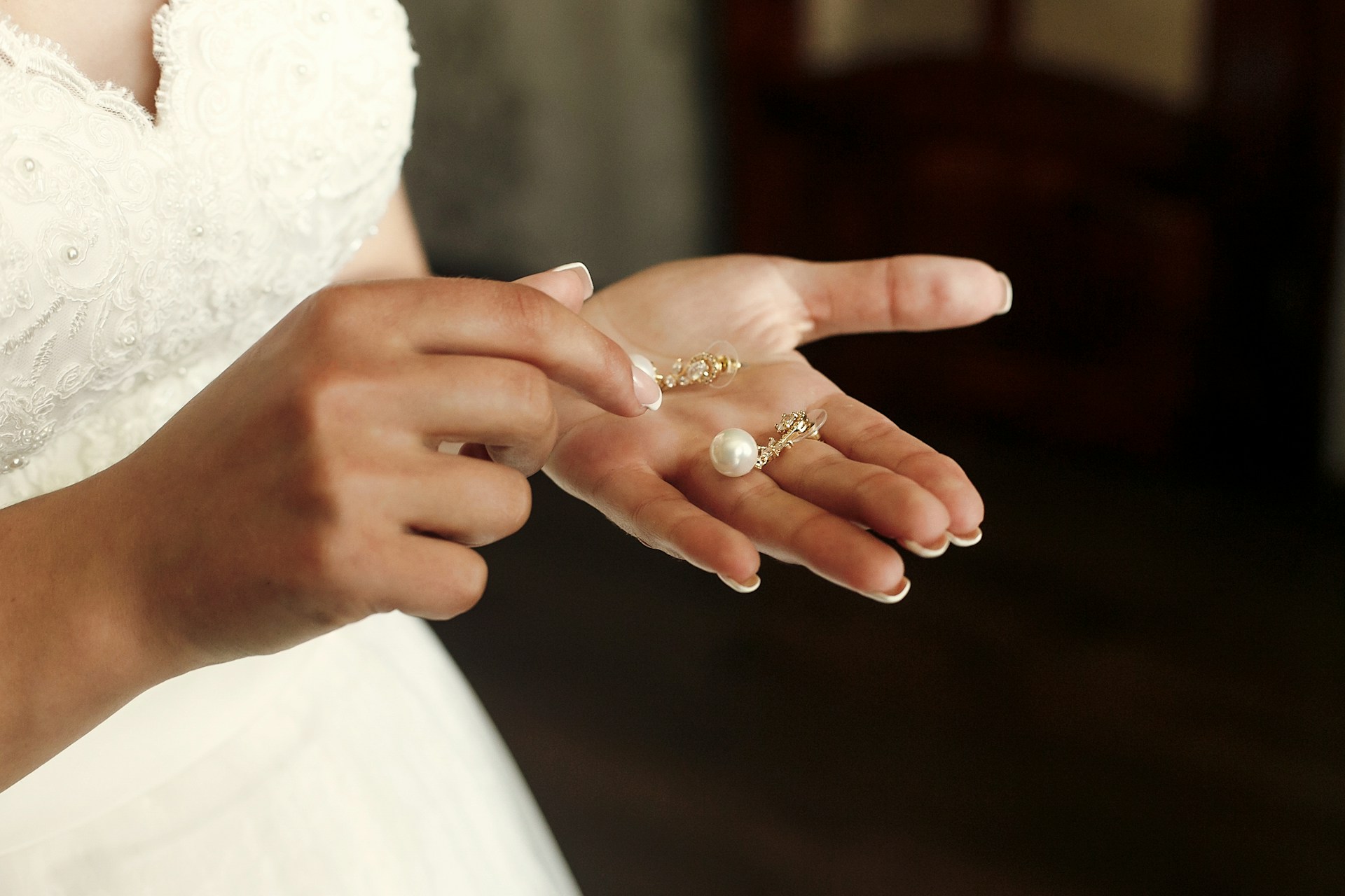 A bride in a white lace wedding gown holds a pair of delicate pearl earrings in her hands, preparing for her special day.
