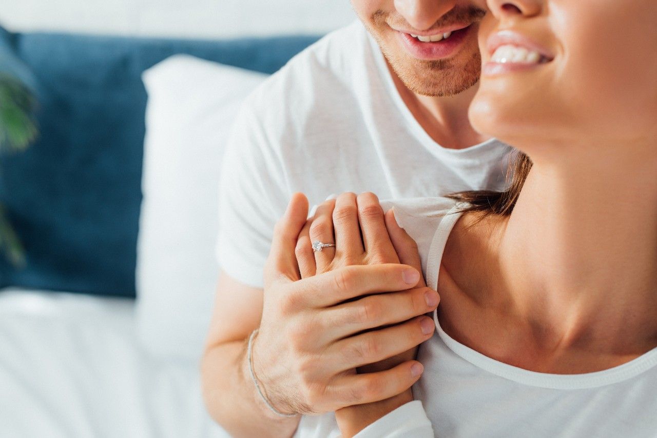 A happy couple in white shirts, with the woman's solitaire engagement ring prominently displayed as they hold hands.