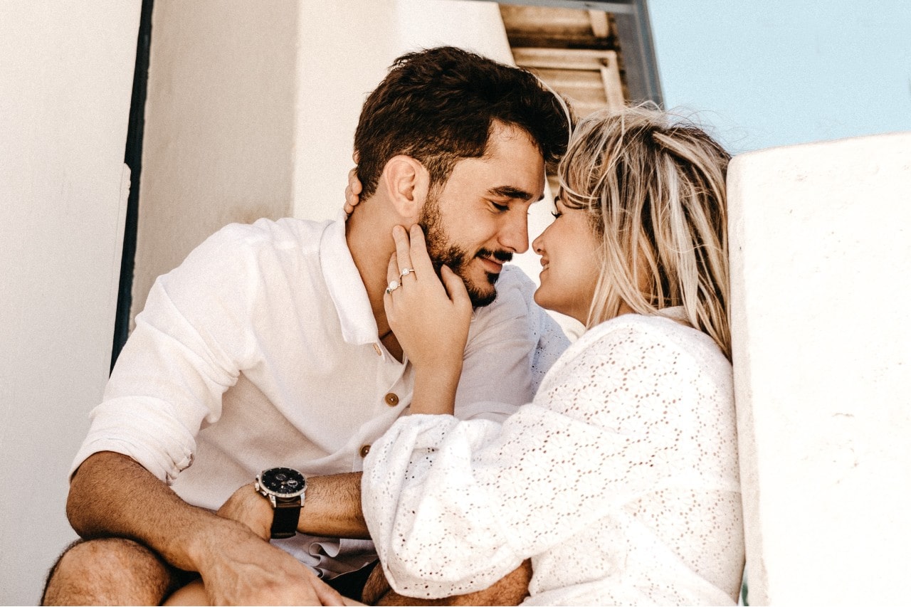 A couple dressed in white, gazing into each other's eyes with loving smiles, as the woman gently touches the man's face, her engagement ring prominently visible.
