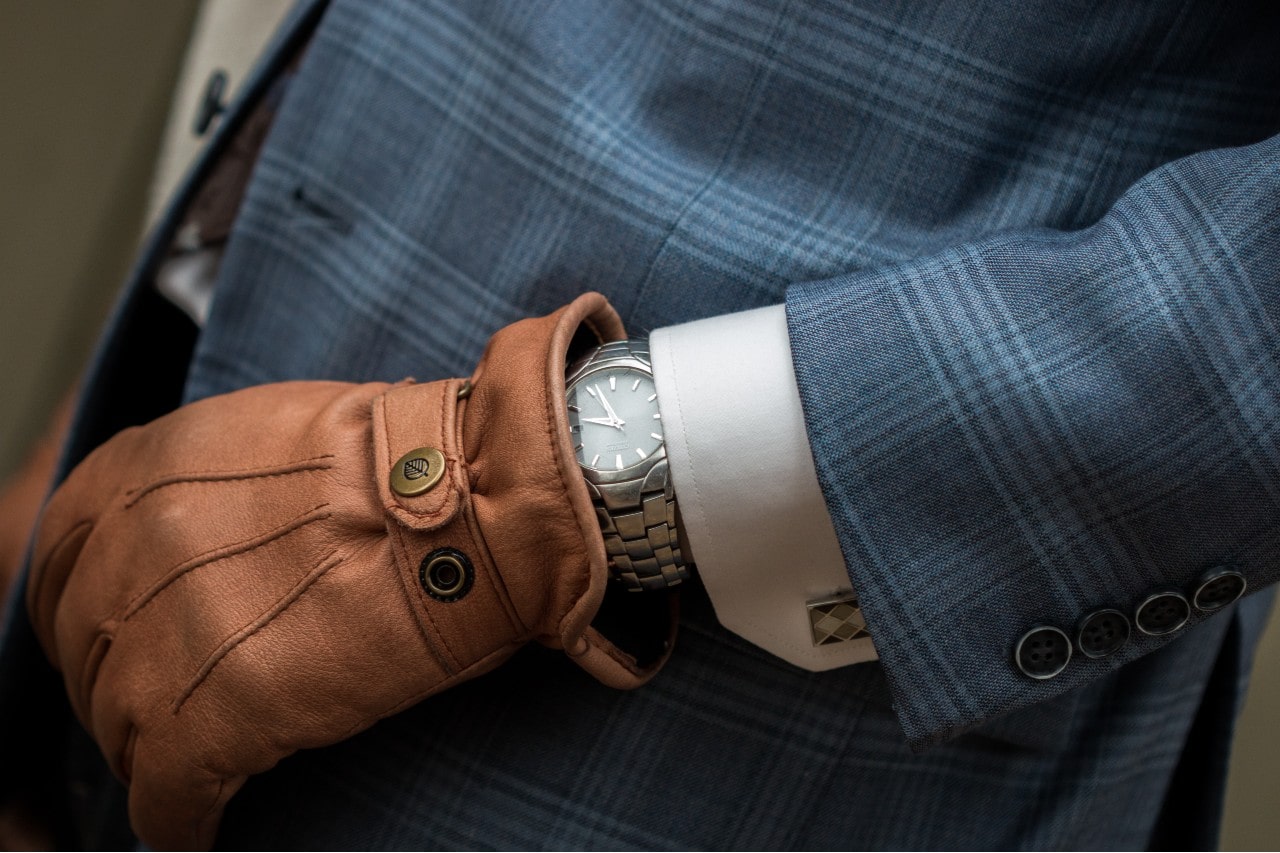 A close-up of a man's wrist wearing a silver wristwatch with a minimalist white dial, peeking out from under a blue plaid suit jacket and white dress shirt. The man is also wearing a brown leather driving glove.