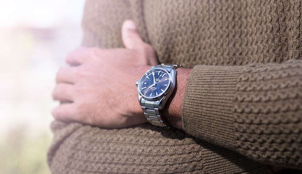 A close-up shot of a man wearing a textured brown sweater, with a stainless steel wristwatch featuring a dark blue dial, Roman numerals, and a date display.