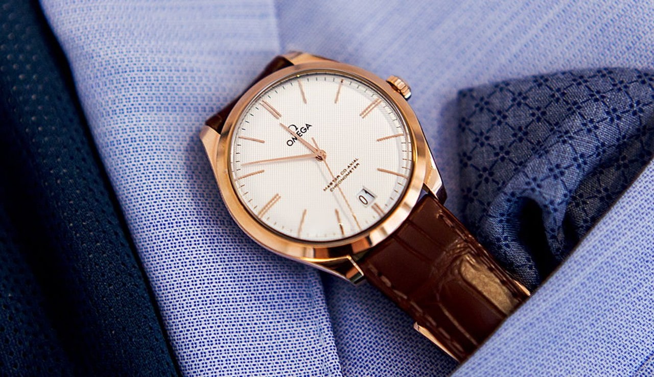 A close-up of a rose gold Omega watch with a white textured face, brown leather strap, and date display, placed on a violet textured suit with a patterned pocket square.