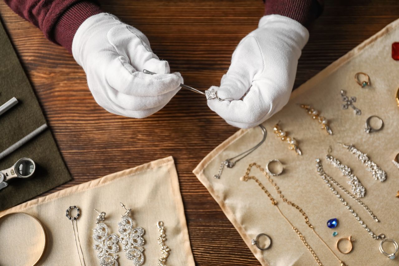 A jeweler wearing white gloves inspects a diamond ring using tweezers and various jewelry pieces are displayed on beige cloths.