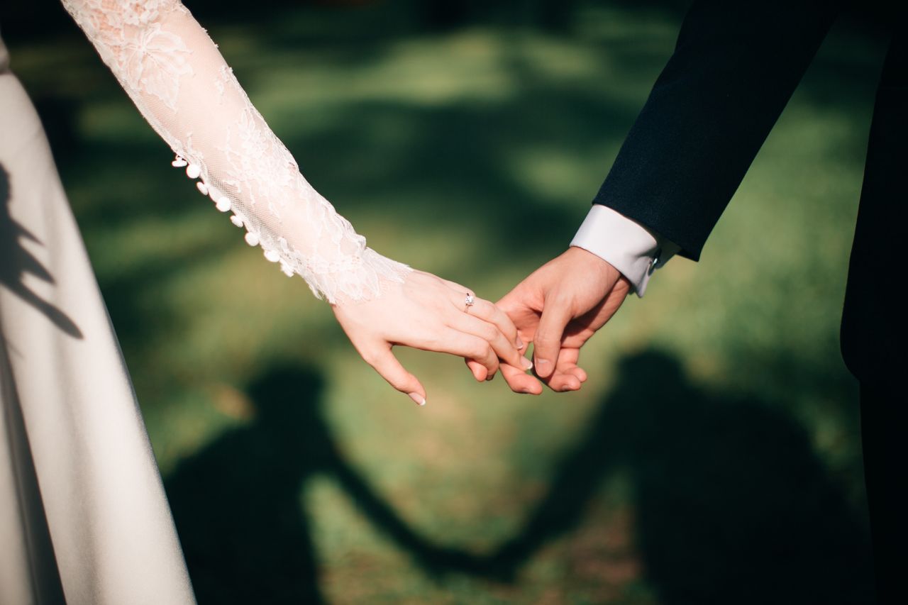 A bride and groom hold hands, their shadows reflect on the grass.