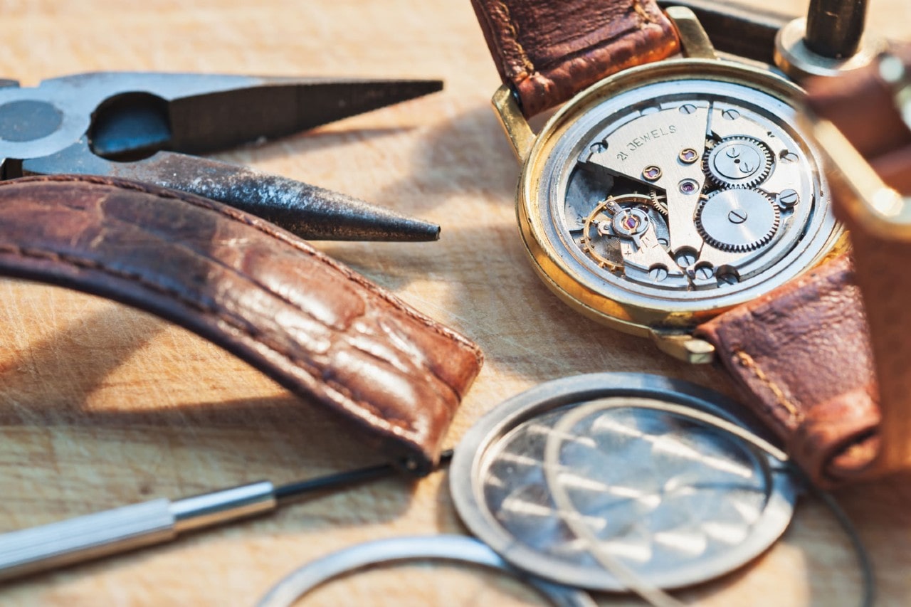 Close-up of a disassembled wristwatch with visible gears and leather strap. Surrounding tools indicate repair work, creating a meticulous and focused atmosphere.