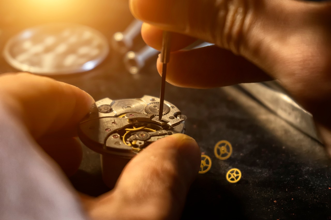 Close-up of hands repairing a watch mechanism with a tiny screwdriver, set against a warm, softly lit background. Loose gears are scattered nearby.