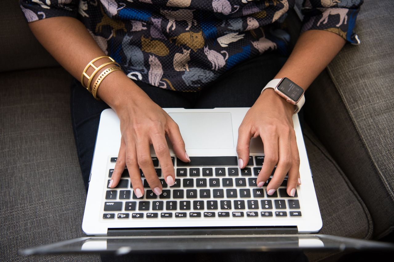 A woman typing on a laptop, her wrists adorned with a smartwatch and yellow gold bracelets.