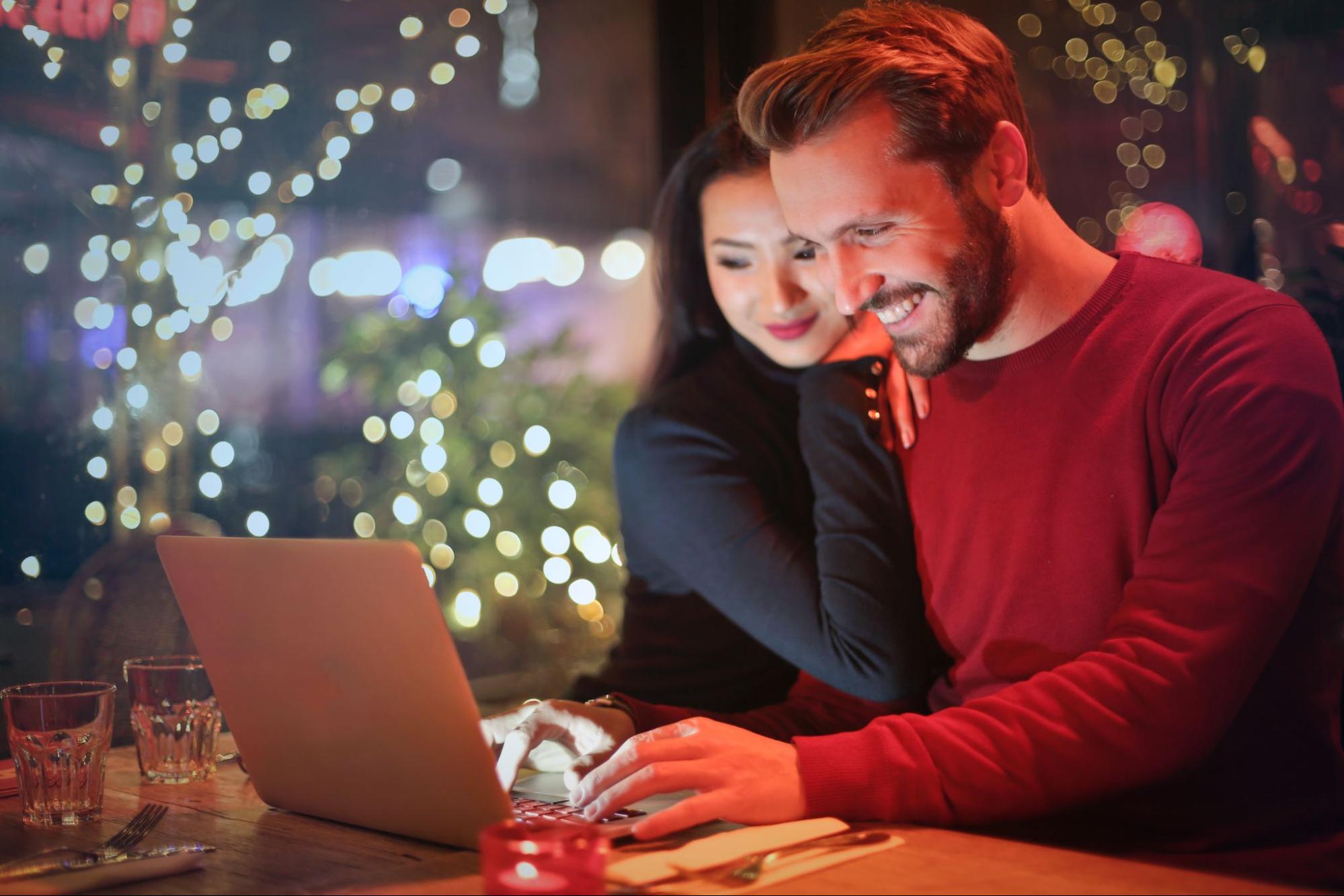 A couple looks joyfully at a laptop screen while sitting in a cafe, surrounded by Christmas tree lighting.