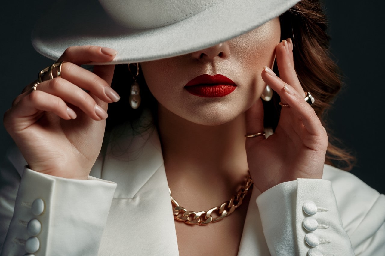 A woman with red lips in a white blazer and white hat, showcasing a yellow gold chain necklace and matching drop earrings and fashion rings.