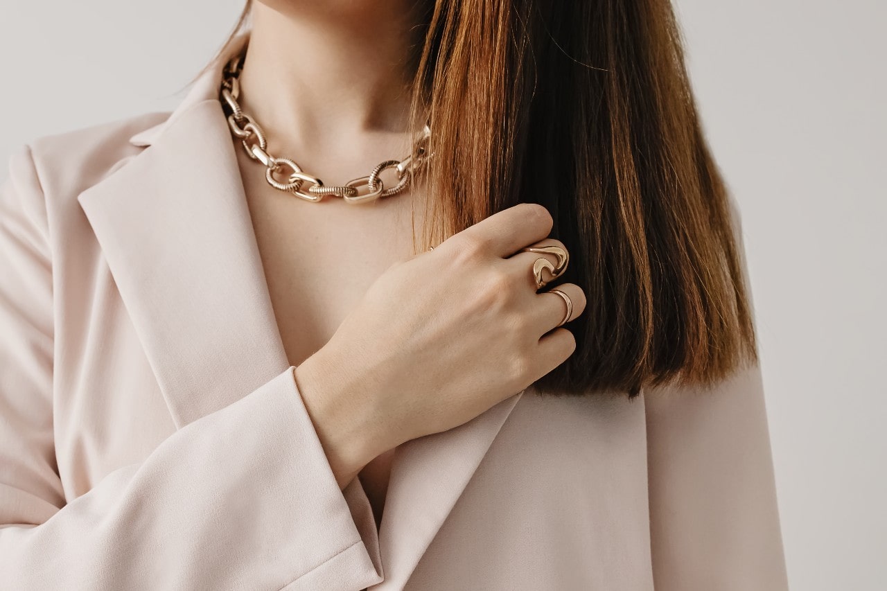 A close-up of a woman&rsquo;s neckline adorned with a yellow gold chain necklace and matching gold rings on her hand.