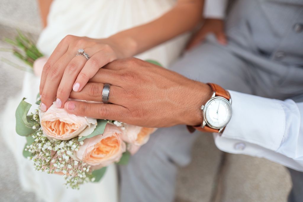 A newly wed couple places their hands on a bouquet of flowers to show off their bridal jewelry