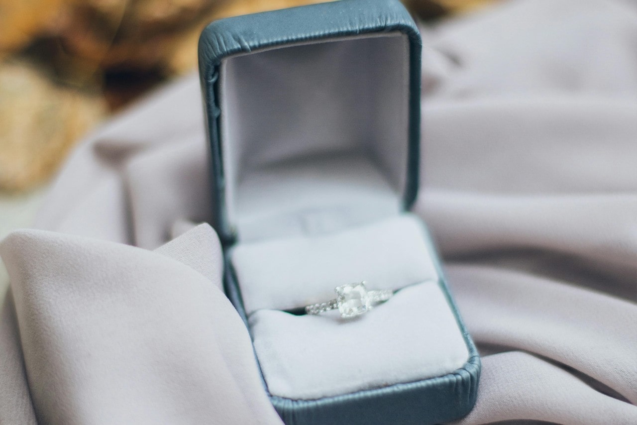 A close-up of an elegant princess cut engagement ring displayed in-box atop a length of white fabric. A close-up of an elegant princess cut engagement ring displayed in-box atop a length of white fabric.