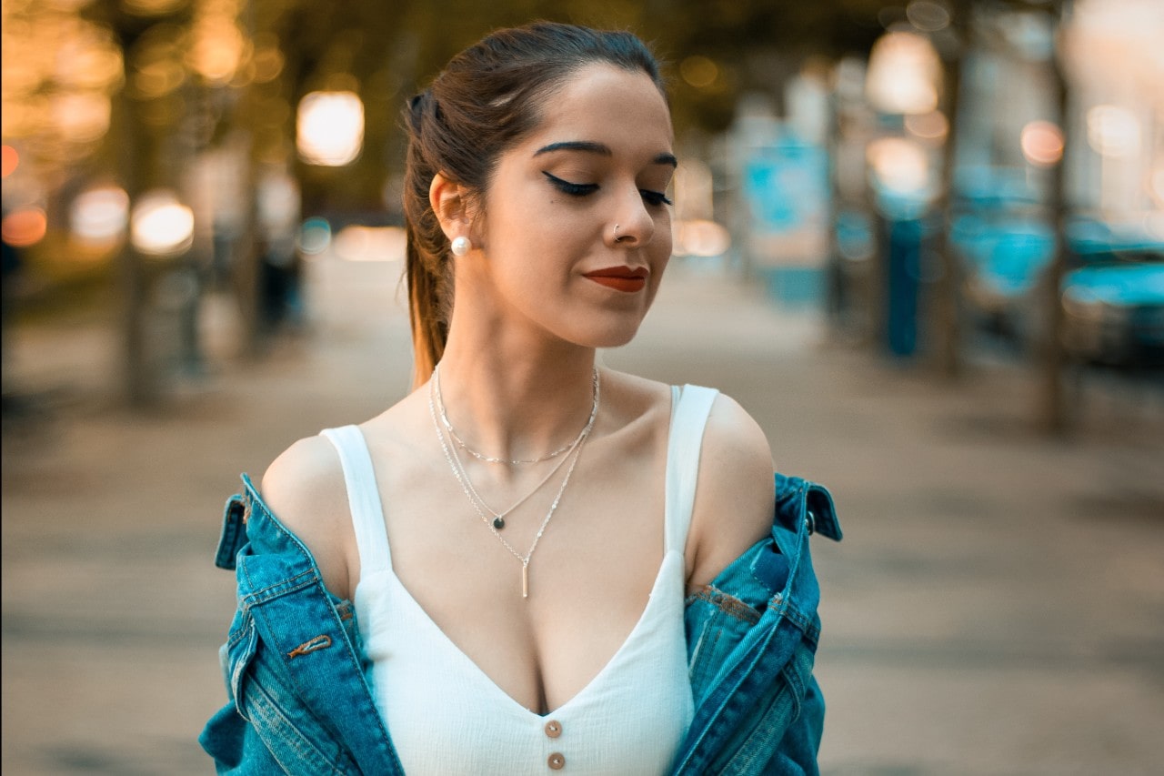 A stylish woman wearing layered necklaces over a white top and denim jacket, posing on a blurred tree-lined street.