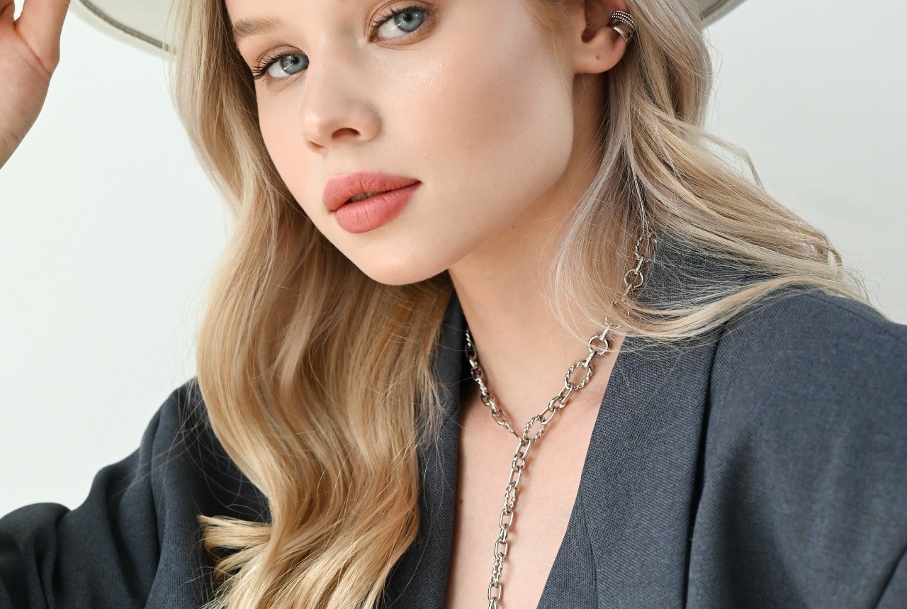 A young woman with long blonde hair wearing a grey blazer and silver chain necklace.