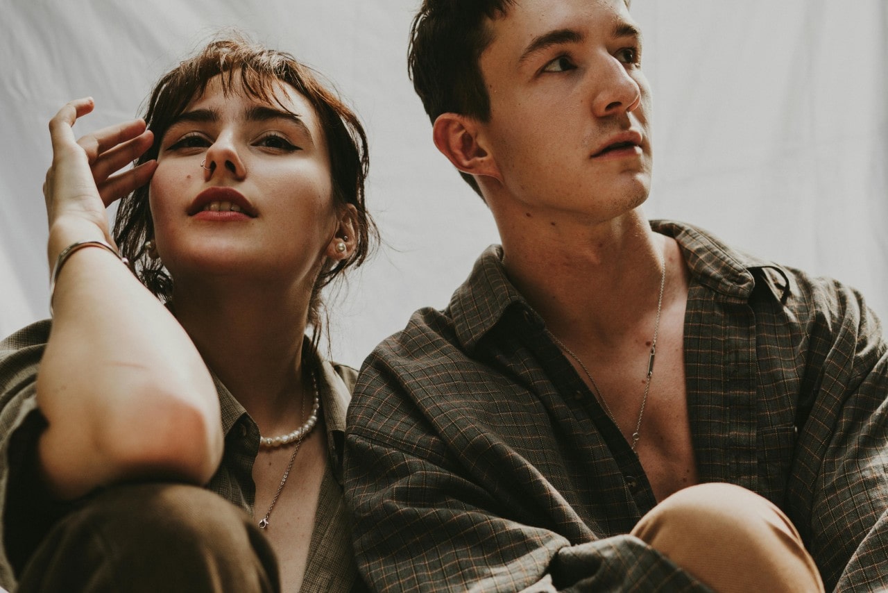 A young woman and man sit closely, gazing in opposite directions, wearing plaid shirts and elegant fashion jewelry in soft light.