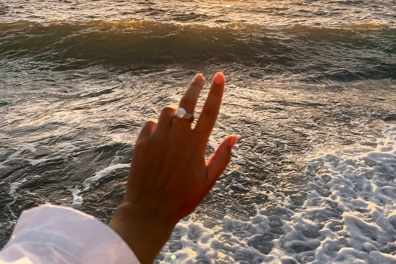 a woman’s hand extended toward the ocean, wearing a large pearl ring