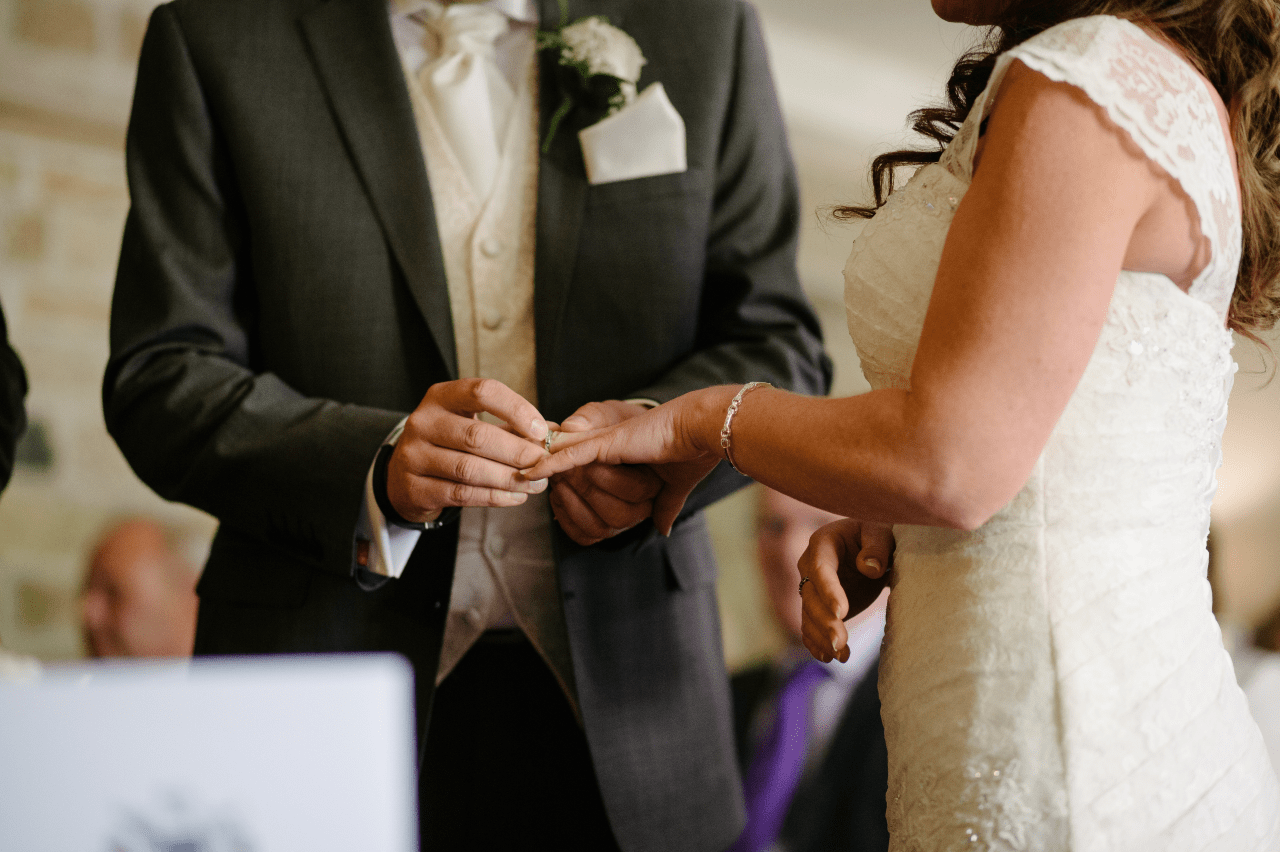 A groom in a dark suit places a wedding ring on the bride’s finger during a wedding ceremony. A groom in a dark suit places a wedding ring on the bride’s finger during a wedding ceremony.