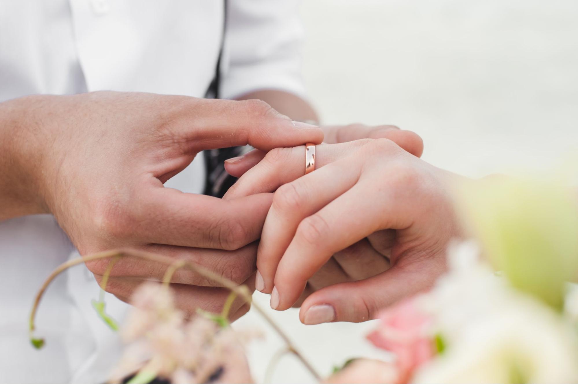 A groom places a gold wedding band on the bride’s finger during a ceremony, surrounded by floral decor. A groom places a gold wedding band on the bride’s finger during a ceremony, surrounded by floral decor.