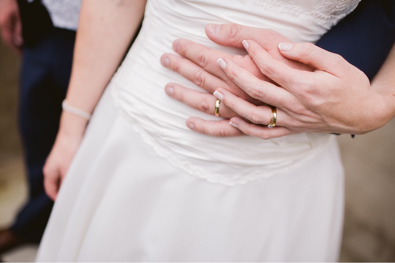 A close-up of a newlywed couple embracing, showcasing their matching gold wedding bands and the bride’s white gown. A close-up of a newlywed couple embracing, showcasing their matching gold wedding bands and the bride’s white gown.