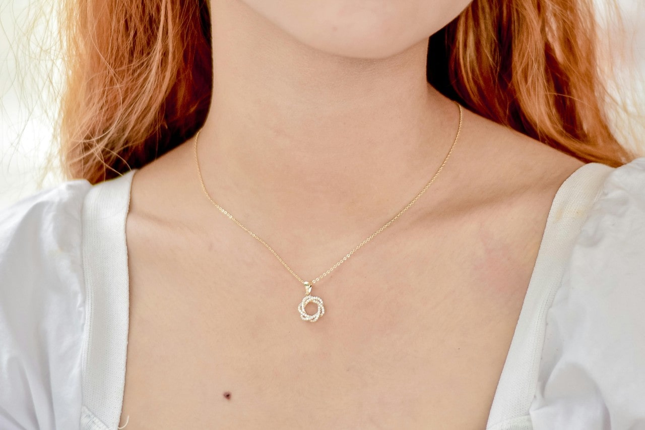 A close-up of a person with red hair wearing a white top, A gold necklace with a small circle pendant adorning their neck.