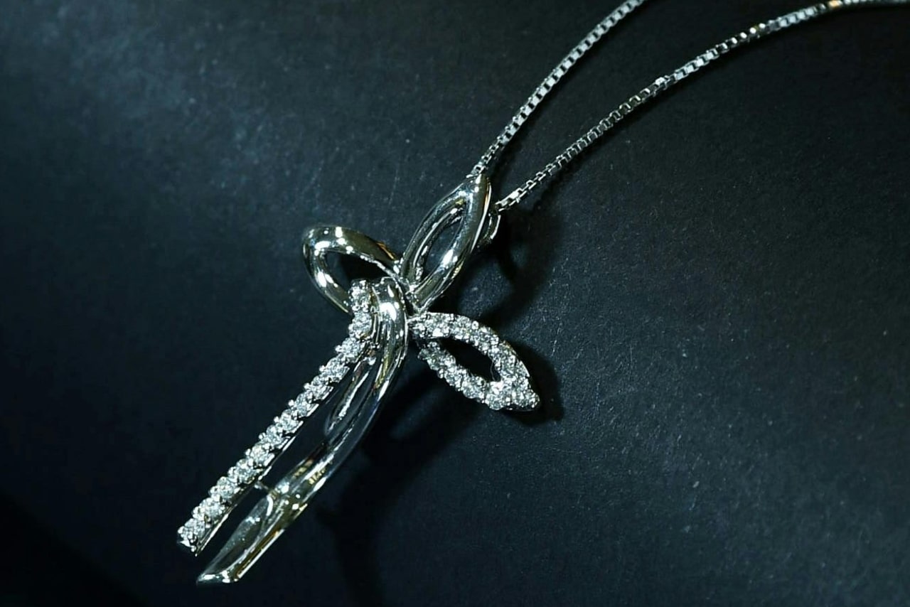 A white gold cross pendant with an intertwined design and sparkling diamond, set against a dark background.