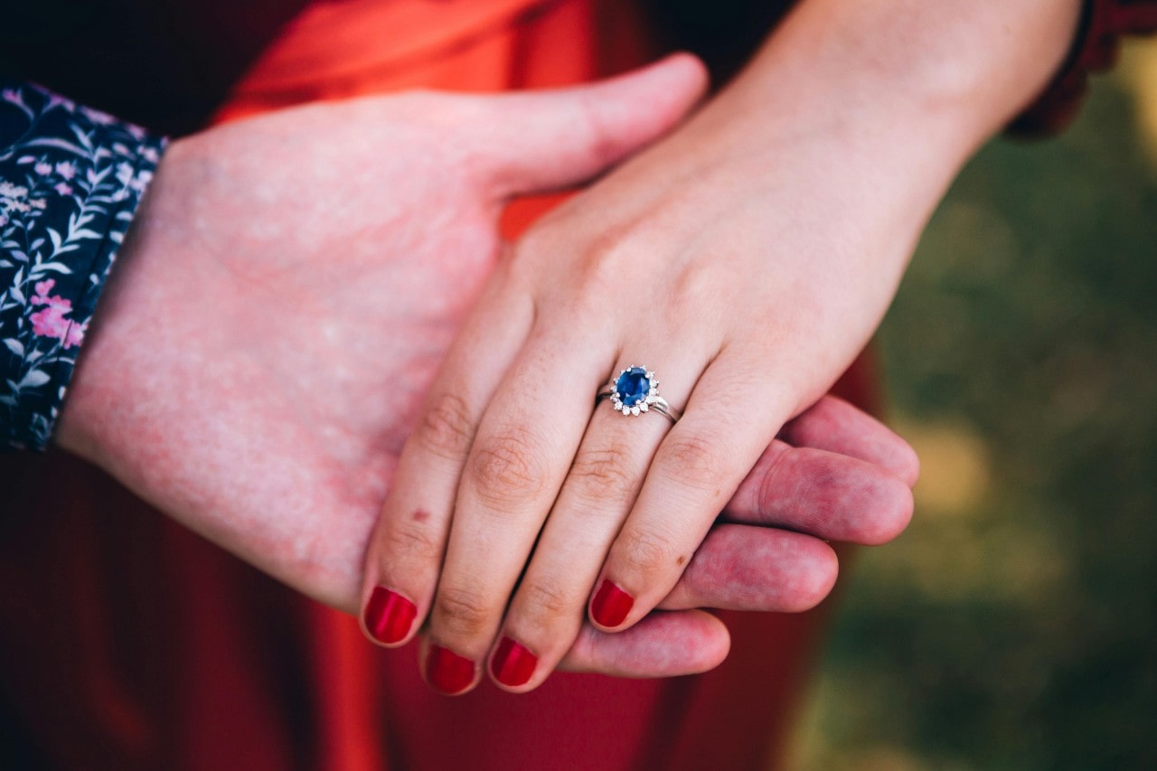 Close-up of a couple’s hand highlighting red nails and a blue sapphire ring of woman against a blurred background.
