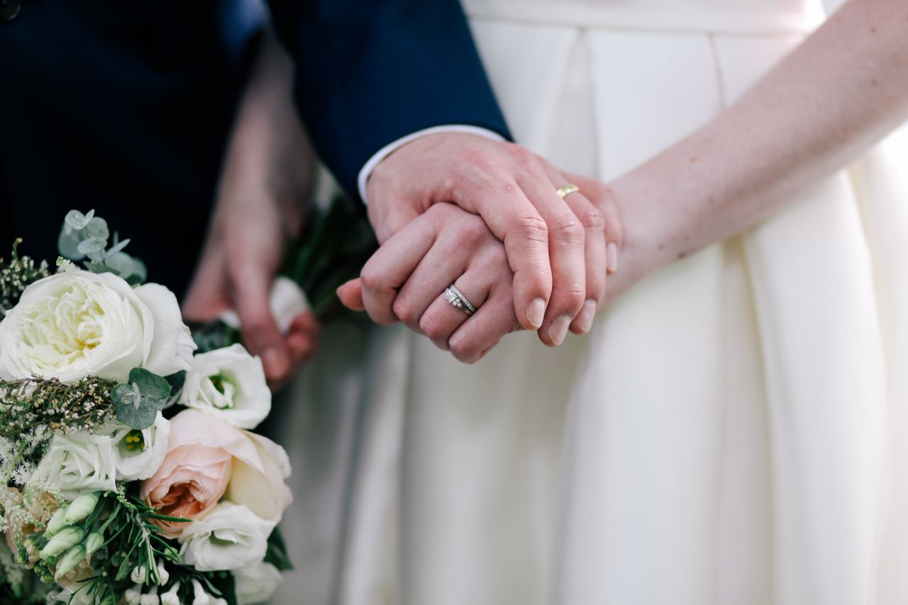 Close-up of a couple holding hands, showcasing a woman’s wedding set and white dress as she holds a bouquet of white and blush roses.
