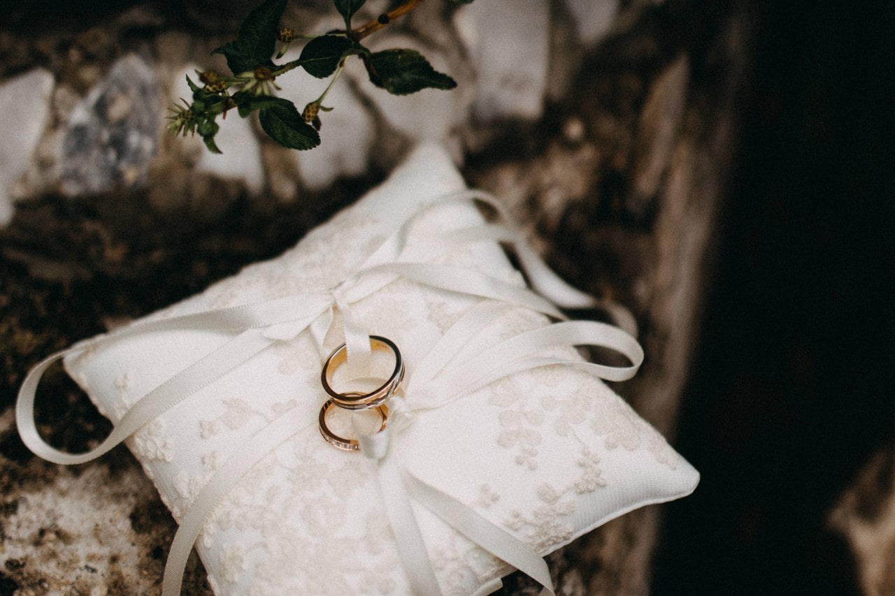 Two yellow gold wedding bands on an ornate white pillow with delicate embroidery and ribbons, placed on a stone surface.