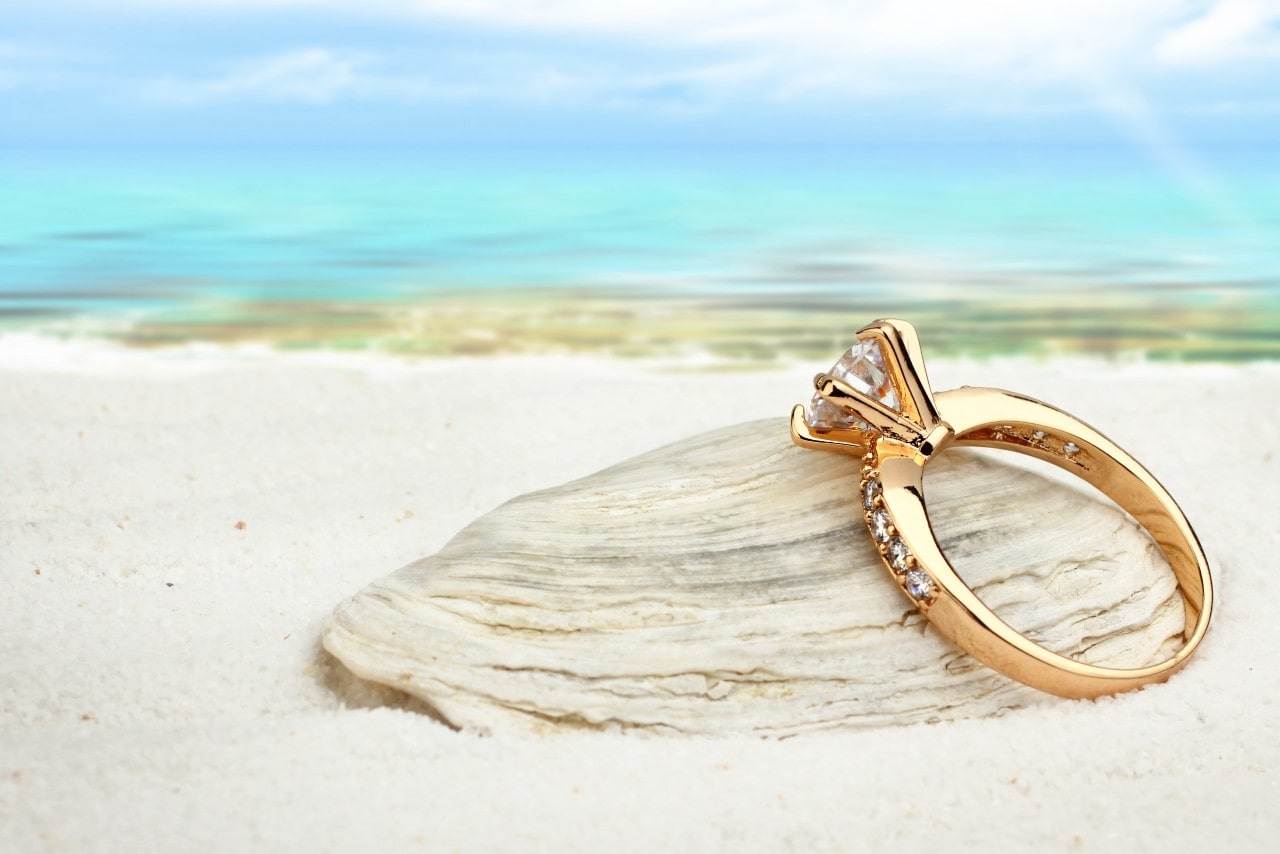 A gold ring with a diamond sits elegantly on a seashell against a serene beach backdrop.