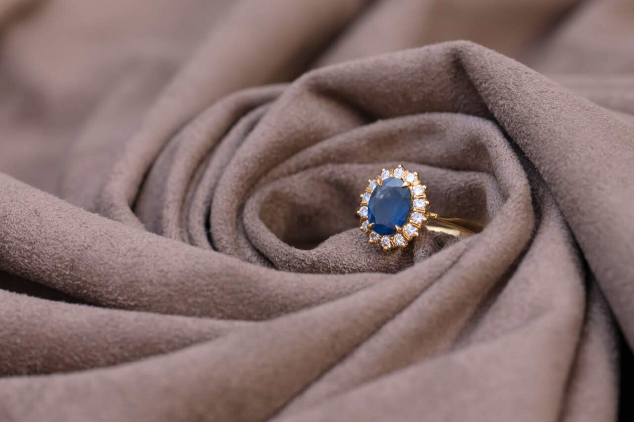 A gold ring with an oval blue gemstone surrounded by small diamonds rests on soft, swirling taupe fabric.