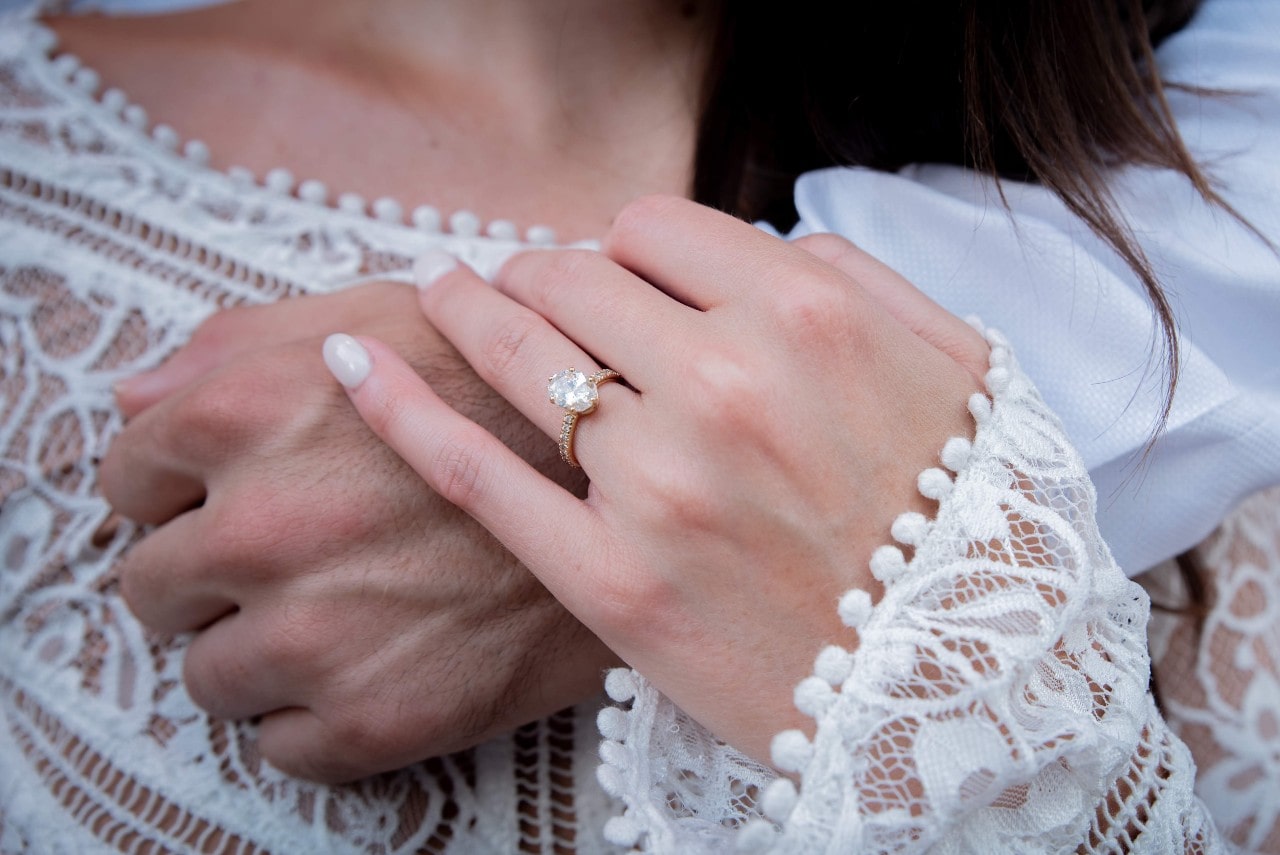 Close-up of a gold engagement ring on a woman’s hand resting on a man’s arm, both in white outfits.